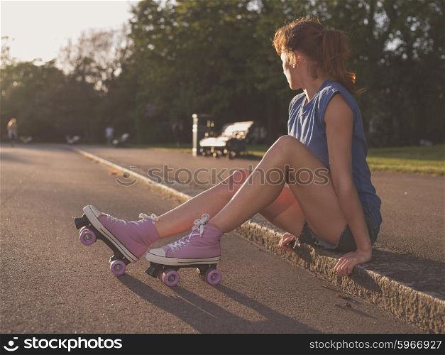 A young woman is sitting on the ground in a park at sunset and she is wearing roller skates
