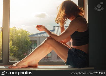 A young woman is sitting on the window sill and looking out on a sunny day