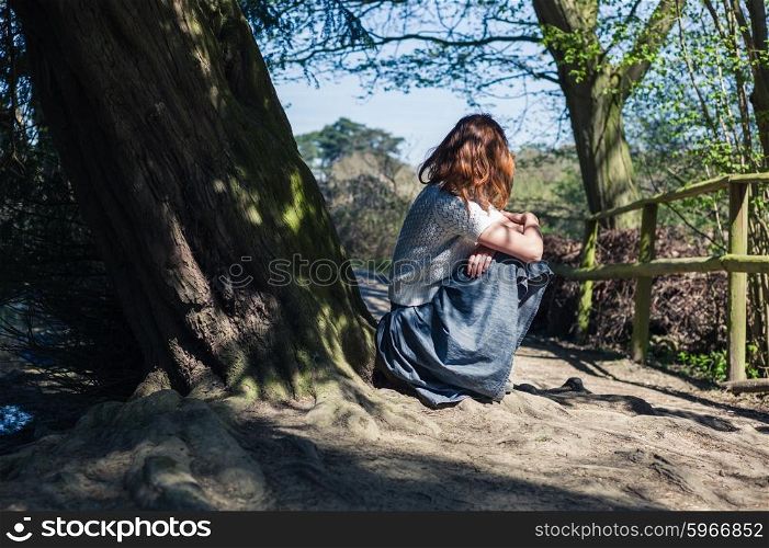 A young woman is sitting under a tree by the water in a forest