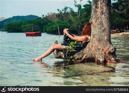 A young woman is sitting up against a tree in the water on a tropical beach