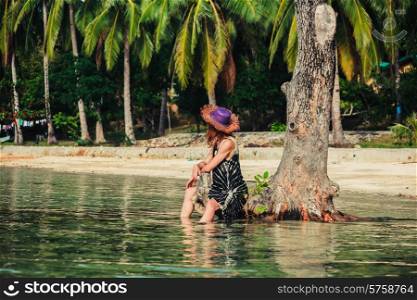A young woman is sitting up against a tree in the water on a tropical beach