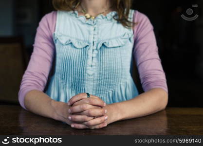 A young woman is sitting with her hands folded at a table in a dining room