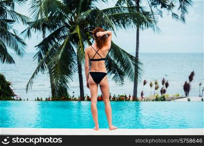 A young woman is standing by a swimming pool with palm trees and the ocean in the background