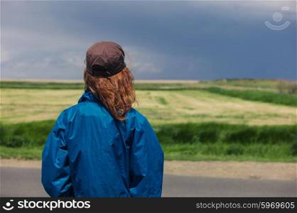 A young woman is standing by the roadside in the country and is looking at a gloomy sky