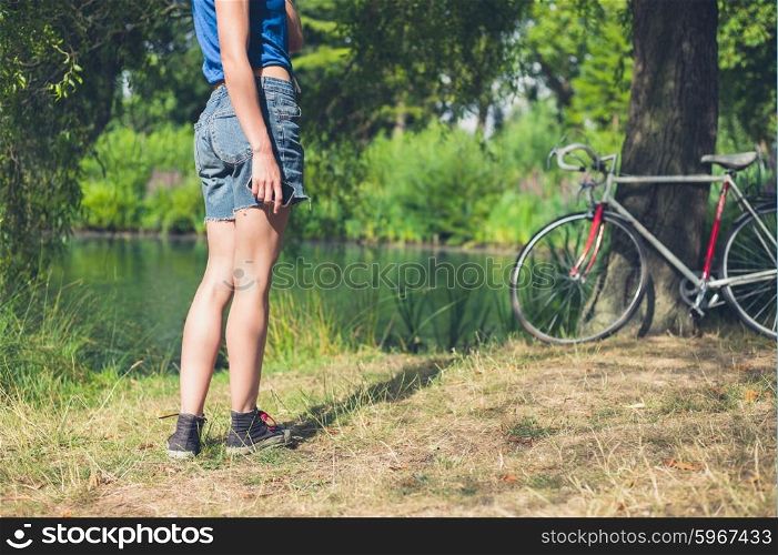 A young woman is standing by the water in a park with her bicycle resting against a tree in the background, she is holding a smart phone in her hand