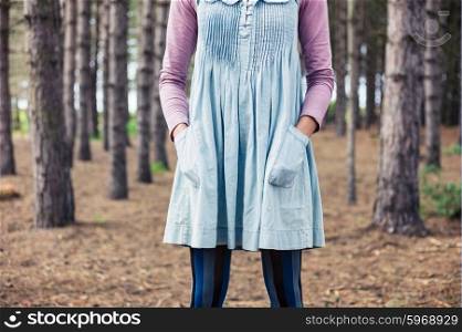 A young woman is standing in the clearing of a forest with her hands in her pockets