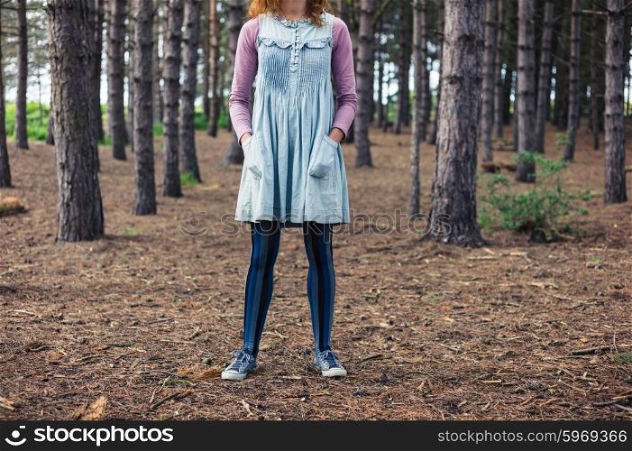 A young woman is standing in the clearing of a forest with her hands in her pockets