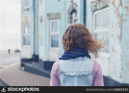 A young woman is standing in the street outside a blue house by the coast on a windy day