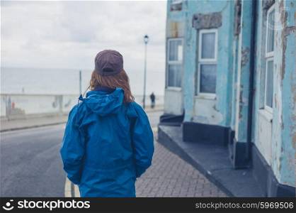 A young woman is standing in the street outside a blue house by the coast on a windy day