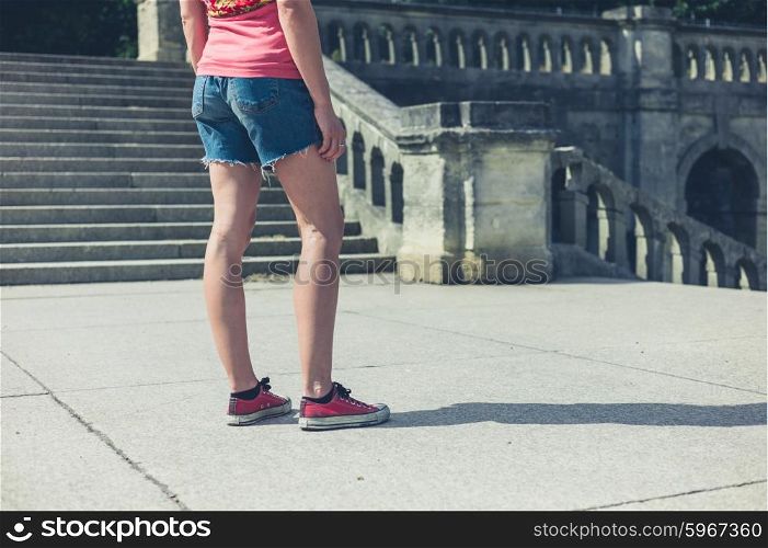 A young woman is standing near the steps of an old Victorian building outside in a park or grand formal garden