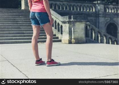 A young woman is standing near the steps of an old Victorian building outside in a park or grand formal garden