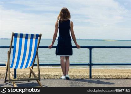 A young woman is standing on a promenade by the seaside with a deck chair and is admiring the sea