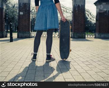 A young woman is standing outside a park by the gates and is holding a skateboard