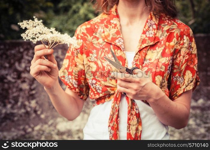 A young woman is standing outside holding a bunch of elderflowers and a pair of scissors