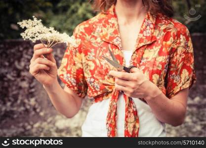 A young woman is standing outside holding a bunch of elderflowers and a pair of scissors