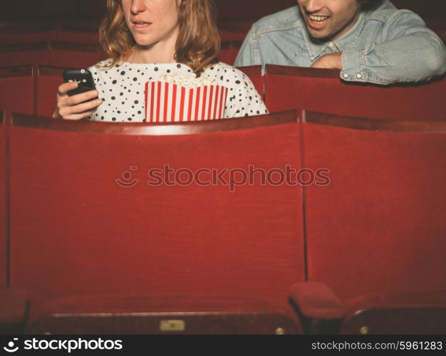 A young woman is using her phone in a movie theater with a man sitting behind her and spying on her