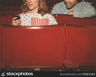 A young woman is using her phone in a movie theater with a man sitting behind her and spying on her