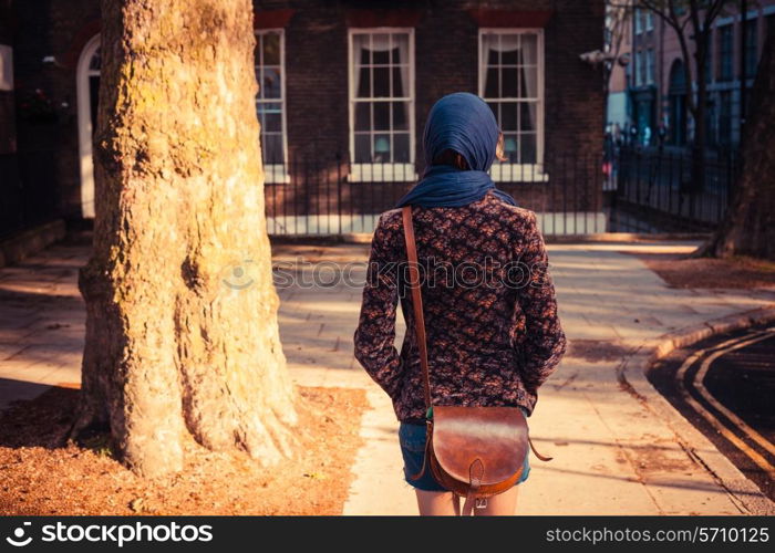 A young woman is walking past a tree and a townhouse in the city on a sunny day