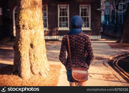 A young woman is walking past a tree and a townhouse in the city on a sunny day