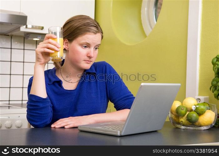 A young woman looks at the computer while drinking juice - surprised by what she reads