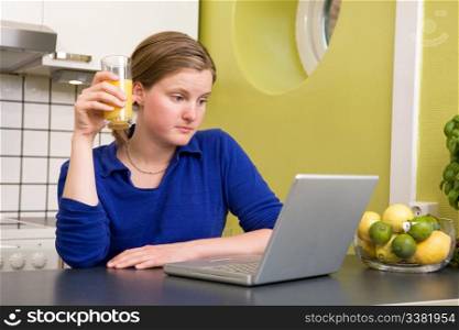 A young woman looks at the computer while drinking juice - surprised by what she reads