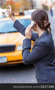 A young woman or businesswoman hailing a yellow Taxi cab while talking on her cell phone in a modern city