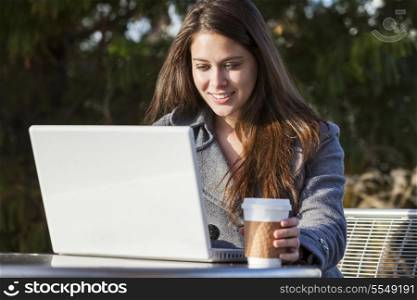 A young woman or girl student using a laptop outside and drinking takeaway coffee