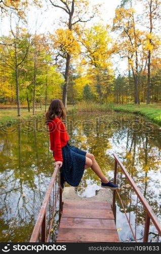 A young woman sits on a bridge near a lake on a sunny day. Beautiful autumn. A young woman sits on a bridge near a lake on a sunny day. Beautiful autumn.