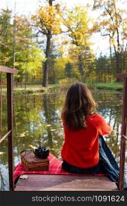 A young woman sits with her back on a bridge near a lake on a sunny day. Beautiful autumn. A young woman sits with her back on a bridge near a lake on a sunny day. Beautiful autumn.