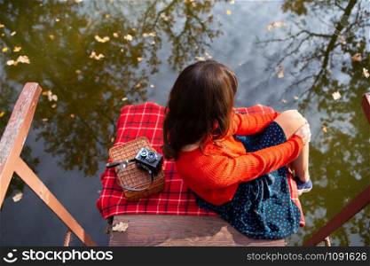 A young woman sits with her back on a bridge near a lake on a sunny day. Plaid, camera, suitcase. A young woman sits with her back on a bridge near a lake on a sunny day. Plaid, camera, suitcase.