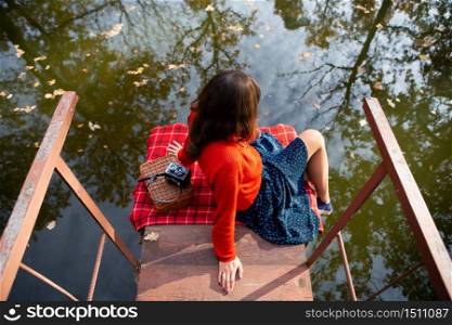 A young woman sits with her back on a bridge near a lake on a sunny day. Plaid, camera, suitcase. A young woman sits with her back on a bridge near a lake on a sunny day. Plaid, camera, suitcase.