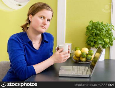 A young woman sitting in the kitchen with a coffee and a computer looking at the camera.