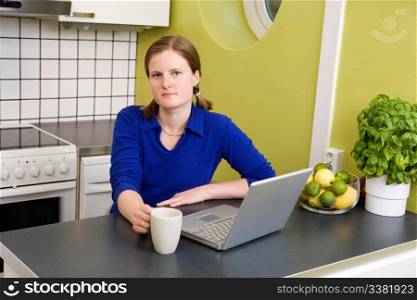 A young woman sitting in the kitchen with a coffee and a computer looking at the camera.