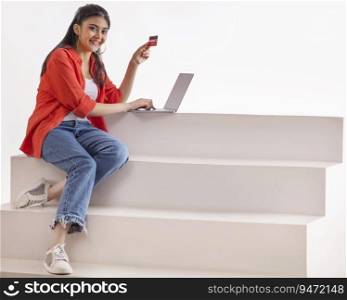 A young woman sitting on stairs making online payment with credit card and laptop.