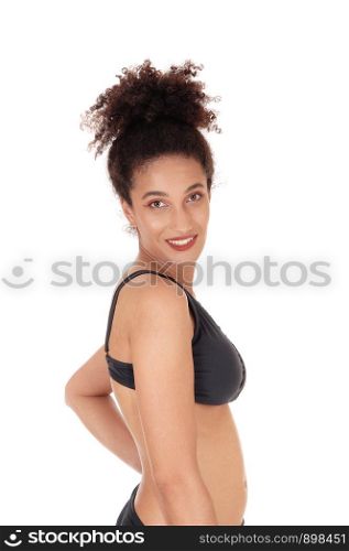 A young woman standing waist up in a black bikini in profile, looking at the camera and smiling, with her curly black hair in a bun, isolatedfor white background