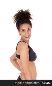 A young woman standing waist up in a black bikini in profile, looking at the camera and smiling, with her curly black hair in a bun, isolatedfor white background