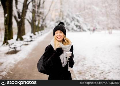 A young woman stands in a winter wonderland, wearing warm clothing and drinking hot coffee to go