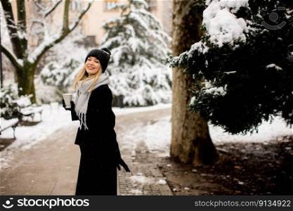 A young woman stands in a winter wonderland, wearing warm clothing and drinking hot coffee to go