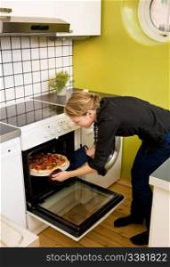 A young woman takes a fresh pizza out of the oven in her apartment kitchen.