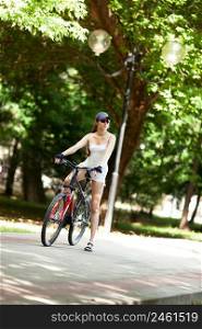 A young woman takes a rest from a bike ride in the park. She’s wearing white sport clothes, cap, her bicycle is red. It’s a sunny summer day.
