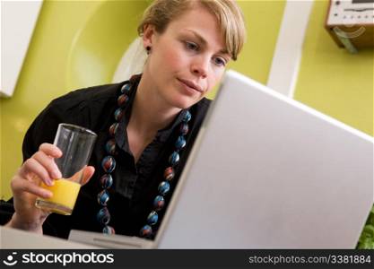 A young woman uses the computer in the kitchen while enjoying a glass of juice. The model is looking at the computer.