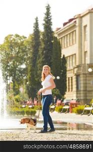 A young woman walking with a beagle dog on a leash near the fountain in the park. Woman walking with a dog