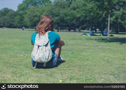 A young woman wearing a backpack is sitting on the grass and relaxing in a park on a summer day