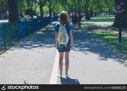 A young woman wearing a bacpack is standig on a path in the park on a summer day