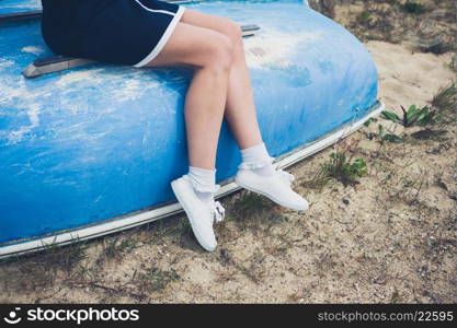 A young woman wearing a blue dress is sitting on an upturned boat on the beach