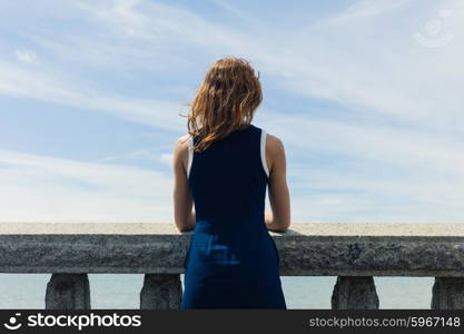 A young woman wearing a blue dress is standing by a wall with concrete balustrades on a promenade and is admiring the seaside on a sunny day