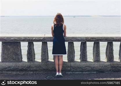 A young woman wearing a blue dress is standing by a wall with concrete balustrades on a promenade and is admiring the seaside on a sunny day