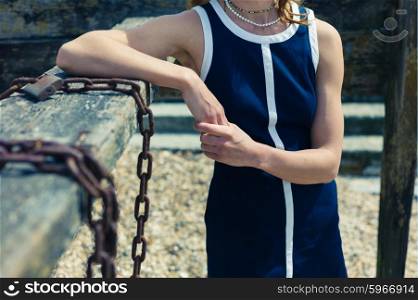A young woman wearing a blue dress is standing on the beach on a sunny day by an old wooden structure with rusty chains hanging from it