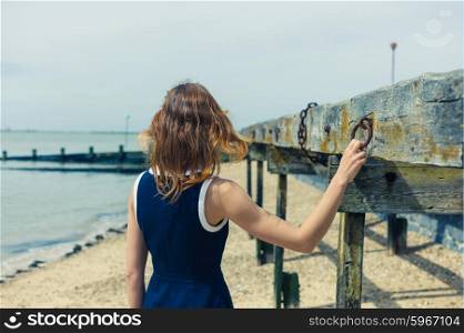 A young woman wearing a blue dress is standing on the beach on a sunny day by an old wooden structure with rusty chains hanging from it