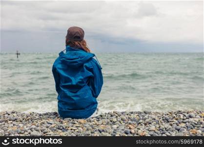 A young woman wearing a blue jacket is sitting on the beach on a windy day